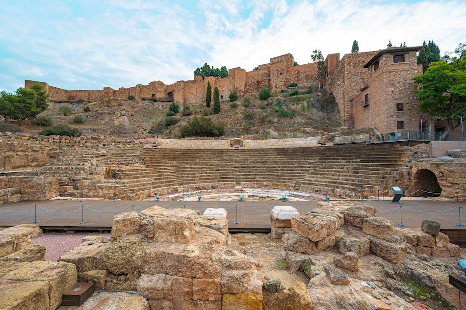 Wide view of the ancient Roman theatre and Alcazaba fortress walls in Malaga, Spain, showing stone seating, wooden stage, and surrounding hillside under a cloudy sky.